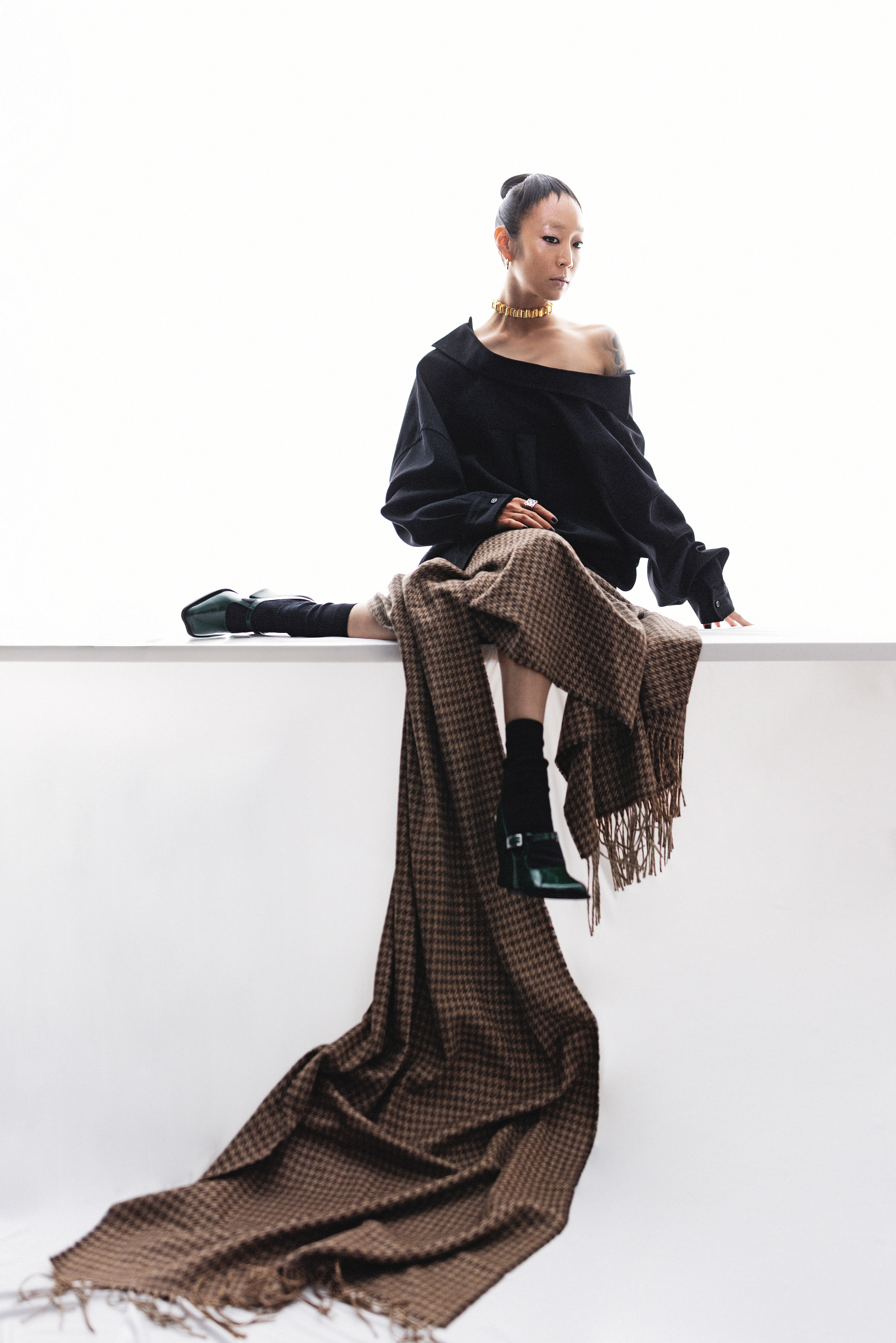 黑色襯衫 NT$34,700 by BOTEGA VENETA；棕色格紋裙 價格店洽 by BALENCIAGA；深綠色方頭高跟鞋 NT$41,000 by LOEWE；金色項鍊 NT$85,400 by GUCCI；棕色格紋流蘇圍巾 價格店洽 by BALENCIAGA；銀色logo戒指 價格店洽 by LOEWE