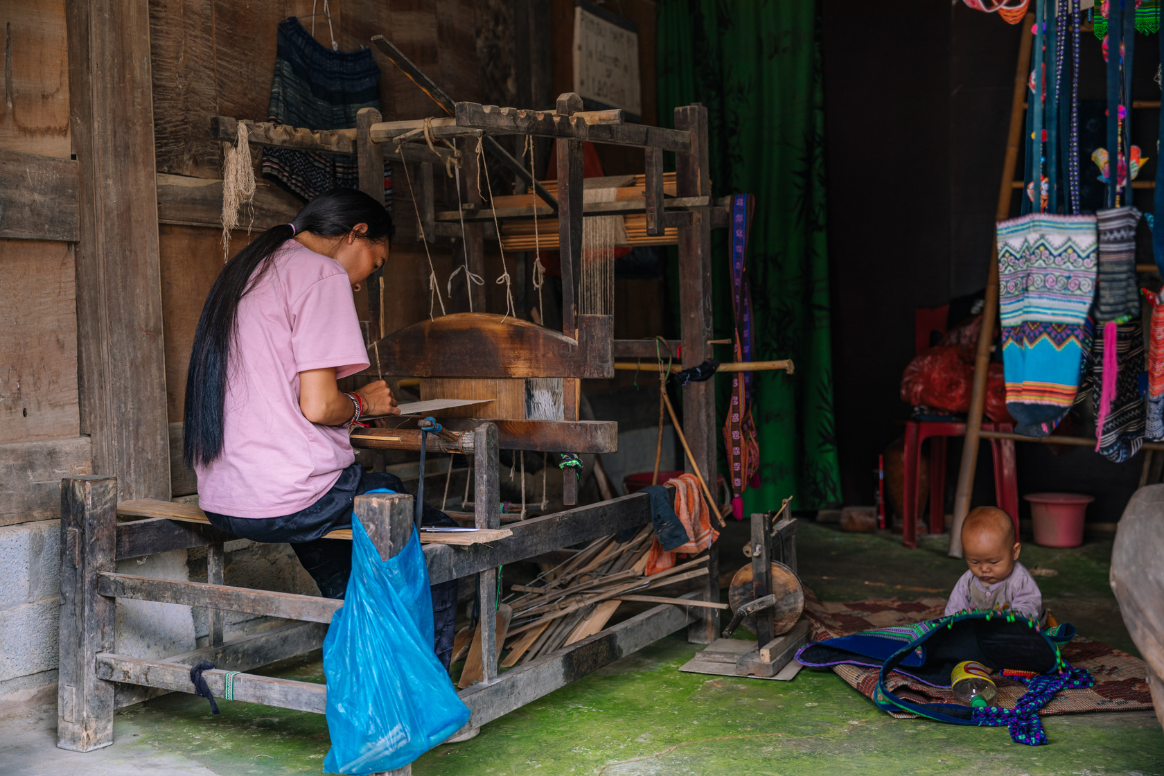 沙壩黑苗部落裡，少女正以傳統手中製作編織物，她的孩子就在一旁地上玩耍。