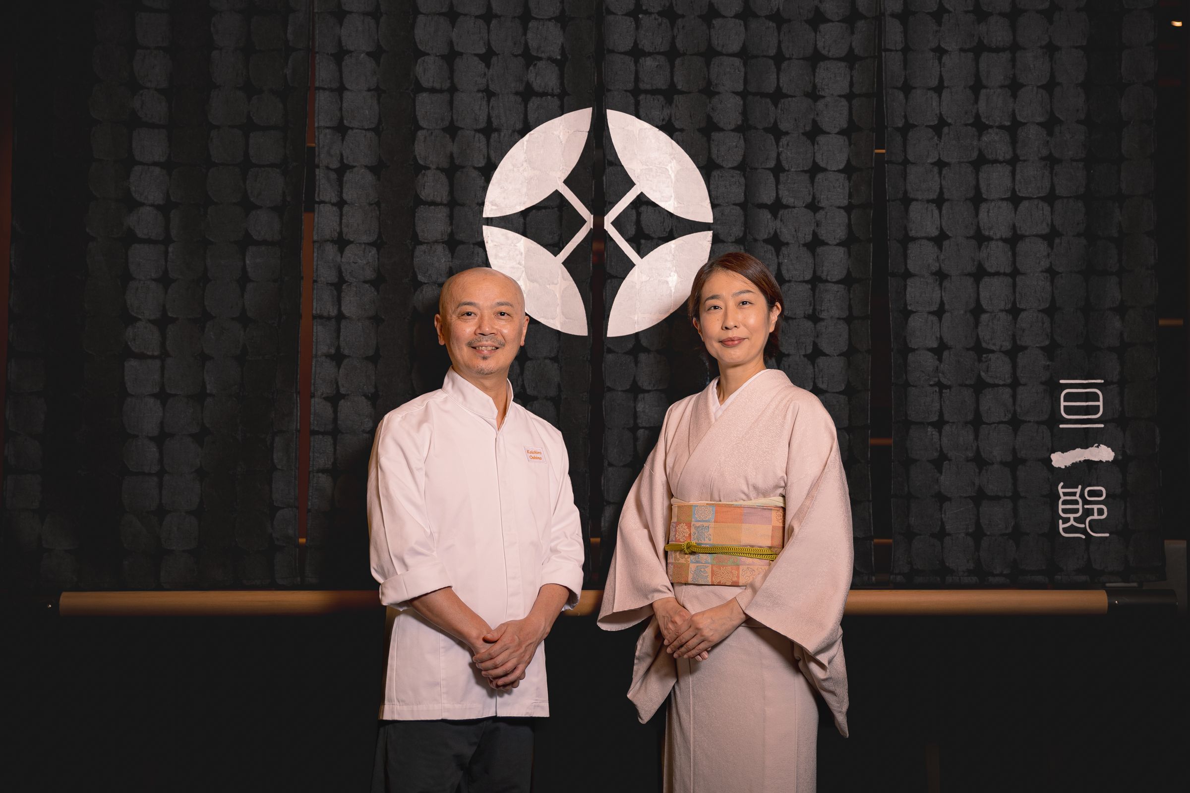 押野亘一郎與素子夫婦從新加坡移居台北,在台北大直打造具有職人精神與款待溫度的高端日料「亘一郎KOICHIRO」,兩人身後是押野家的家徽。(赫士盟提供)