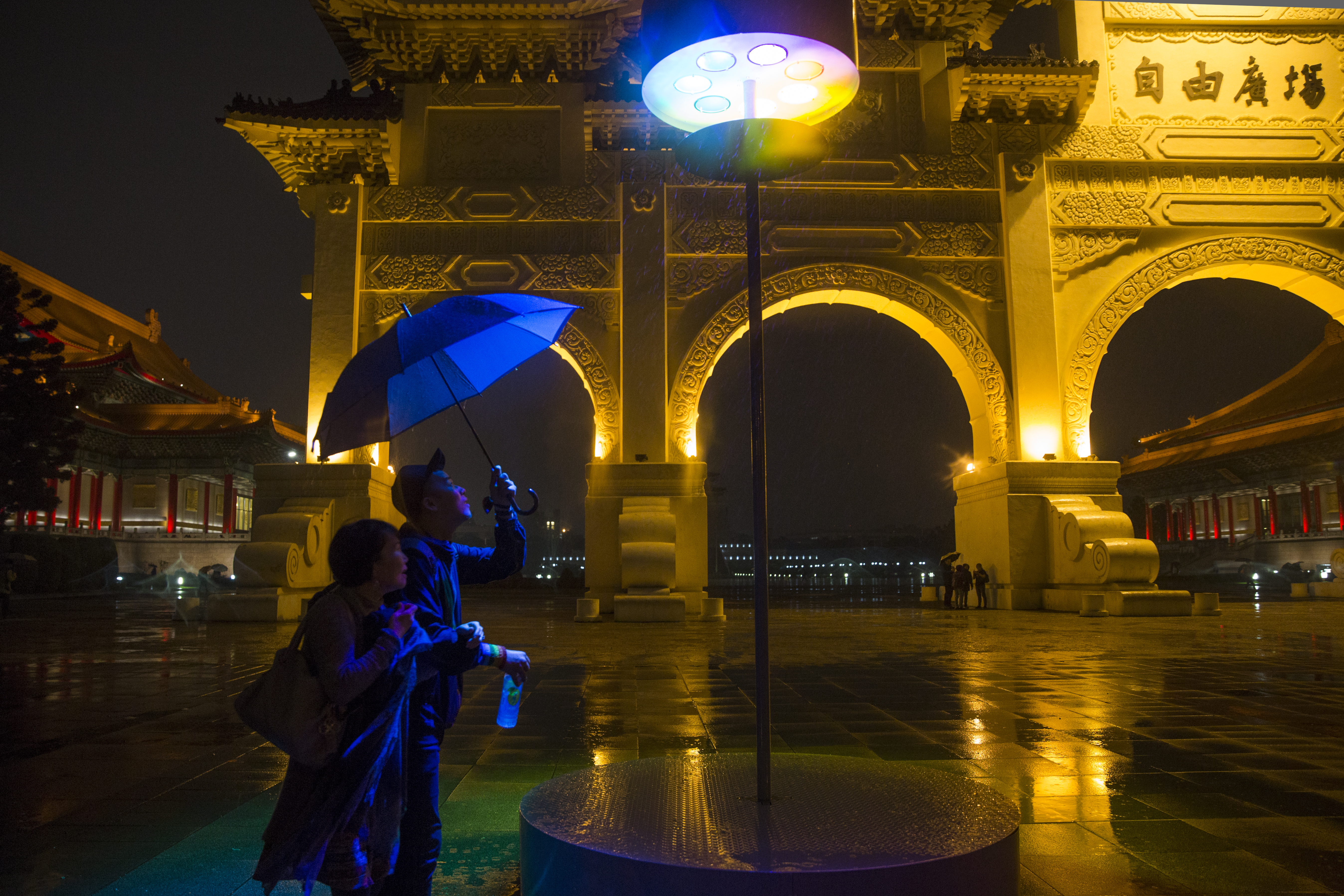 兩人在雨中走過自由廣場，一盞七色的燈光投射在他們身上。