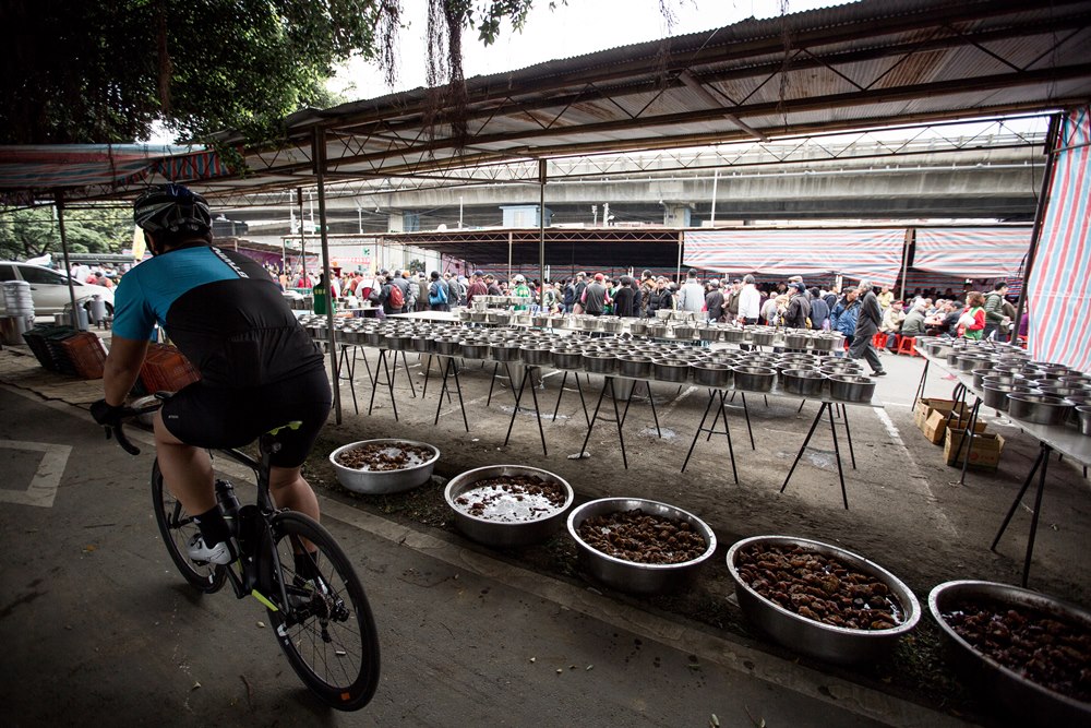 近3年愛心宴遷至萬華河堤外的河濱公園舉辦，辦桌場所旁常有單車騎士經過。