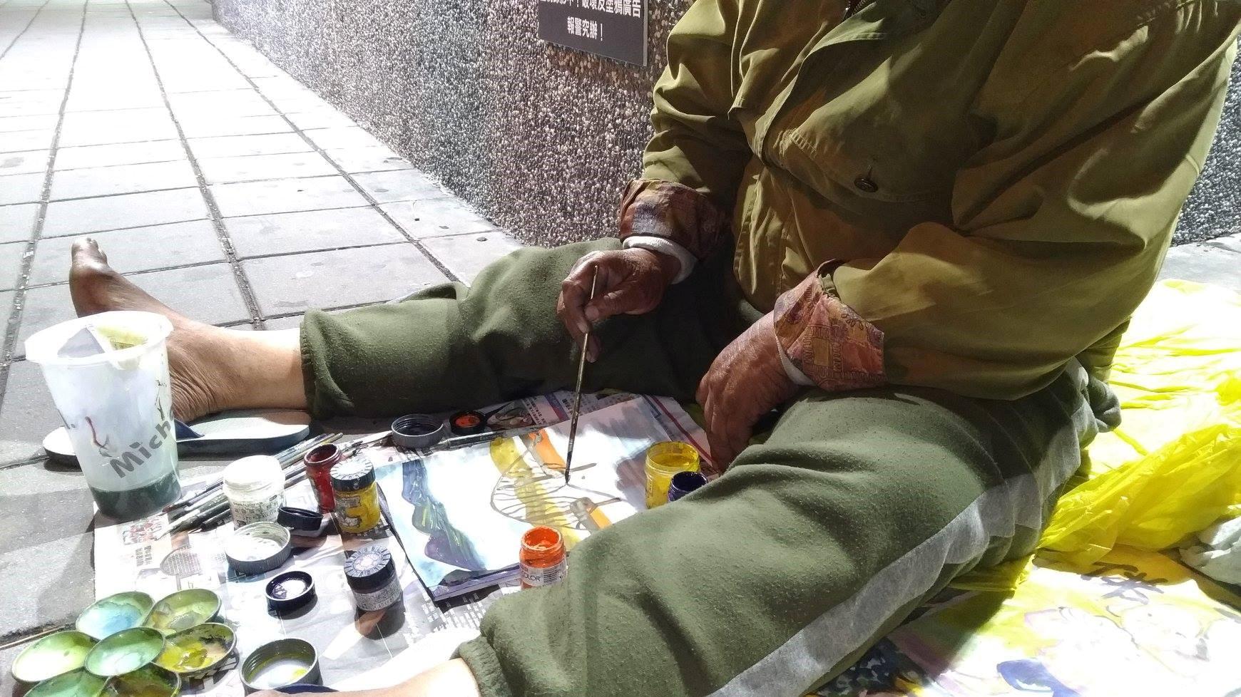 街友阿伯坐在地上作畫。(時代力量三民區議員參選人林于凱提供)