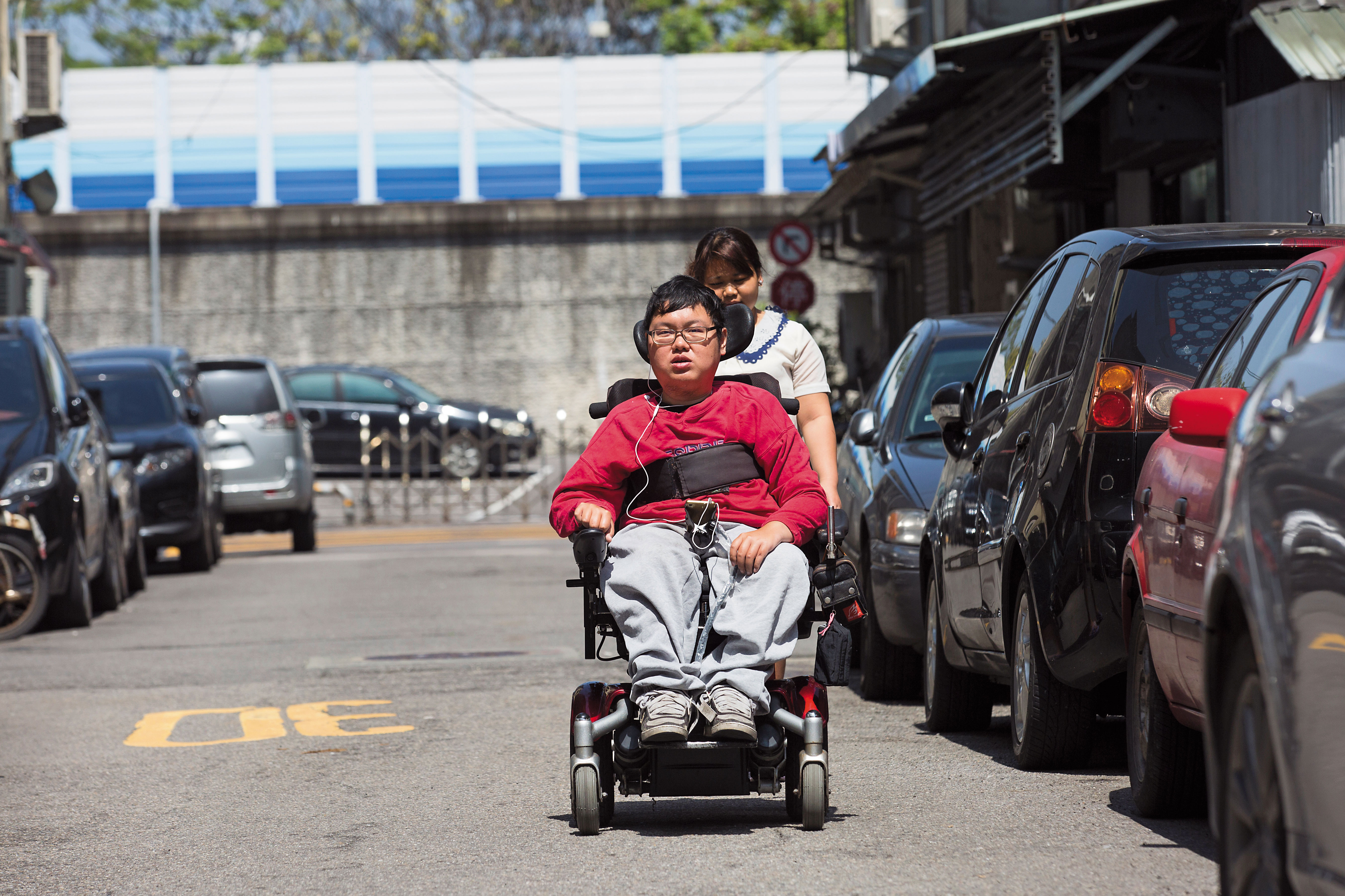 劉于濟（前）是極重度肌肉萎縮症患者，除了推動電動輪椅操控桿，其他都需外傭（後）協助。