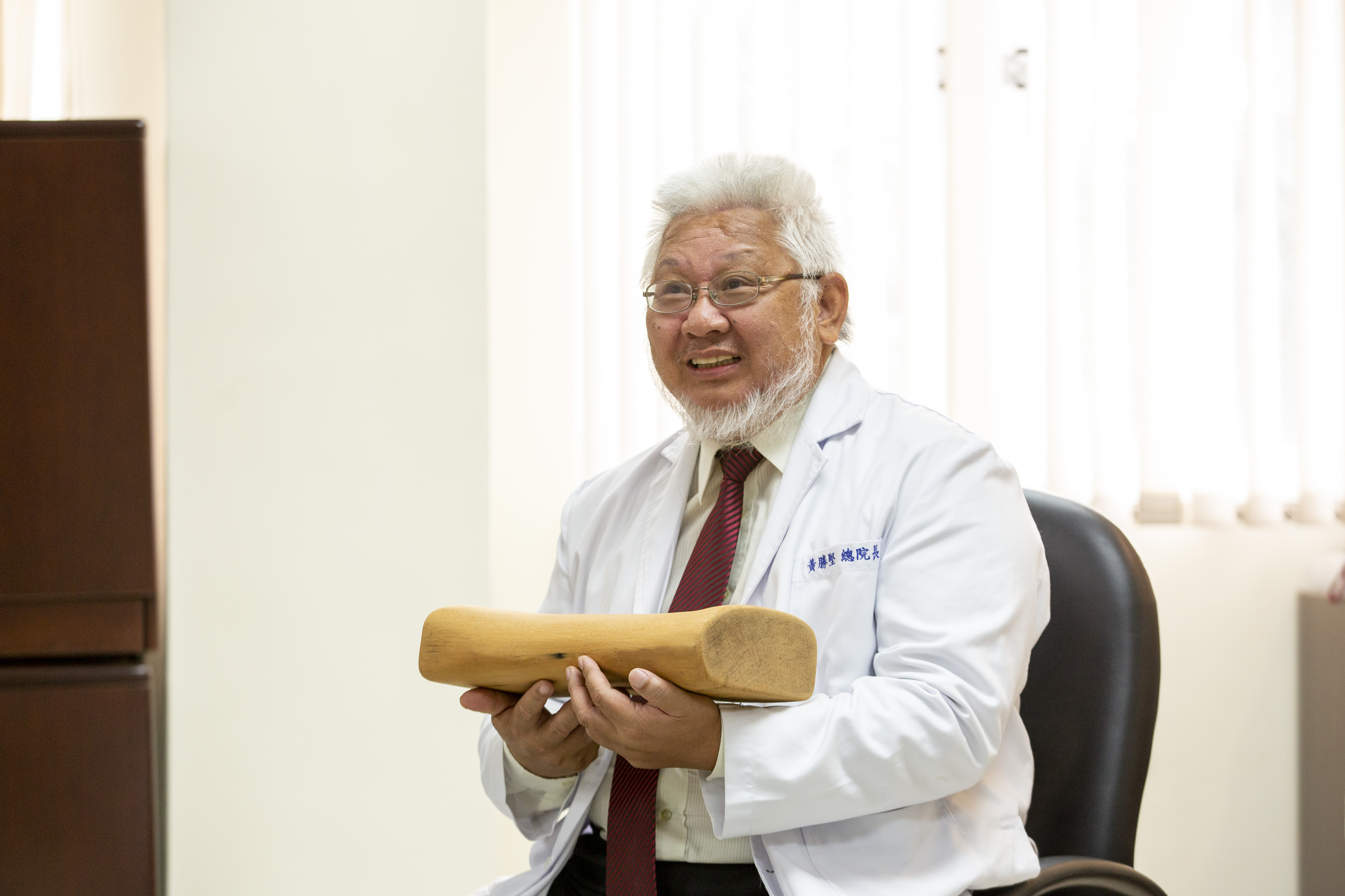 黃勝堅談生死之事直來直往，心態豁達，手中的木枕頭，是一位習慣睡木枕頭的阿嬤過世後，兒子親手雕造贈送，感謝黃勝堅的照顧。