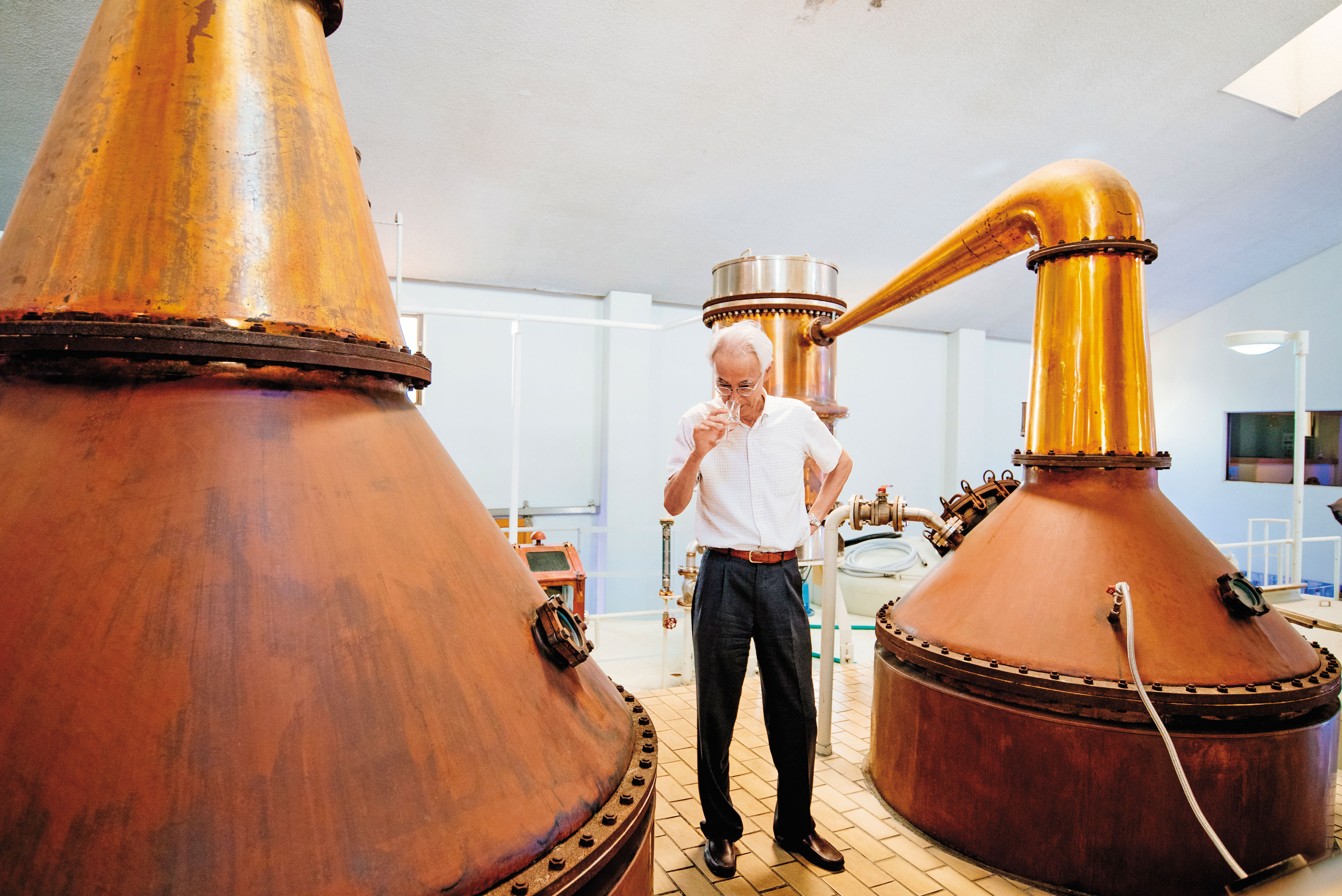 社長平石幹郎站在一對紅銅蒸餾器旁，嗅聞剛蒸餾出來的新酒。左為一次蒸餾器，5,000公升；右為二次蒸餾器，3,000公升。