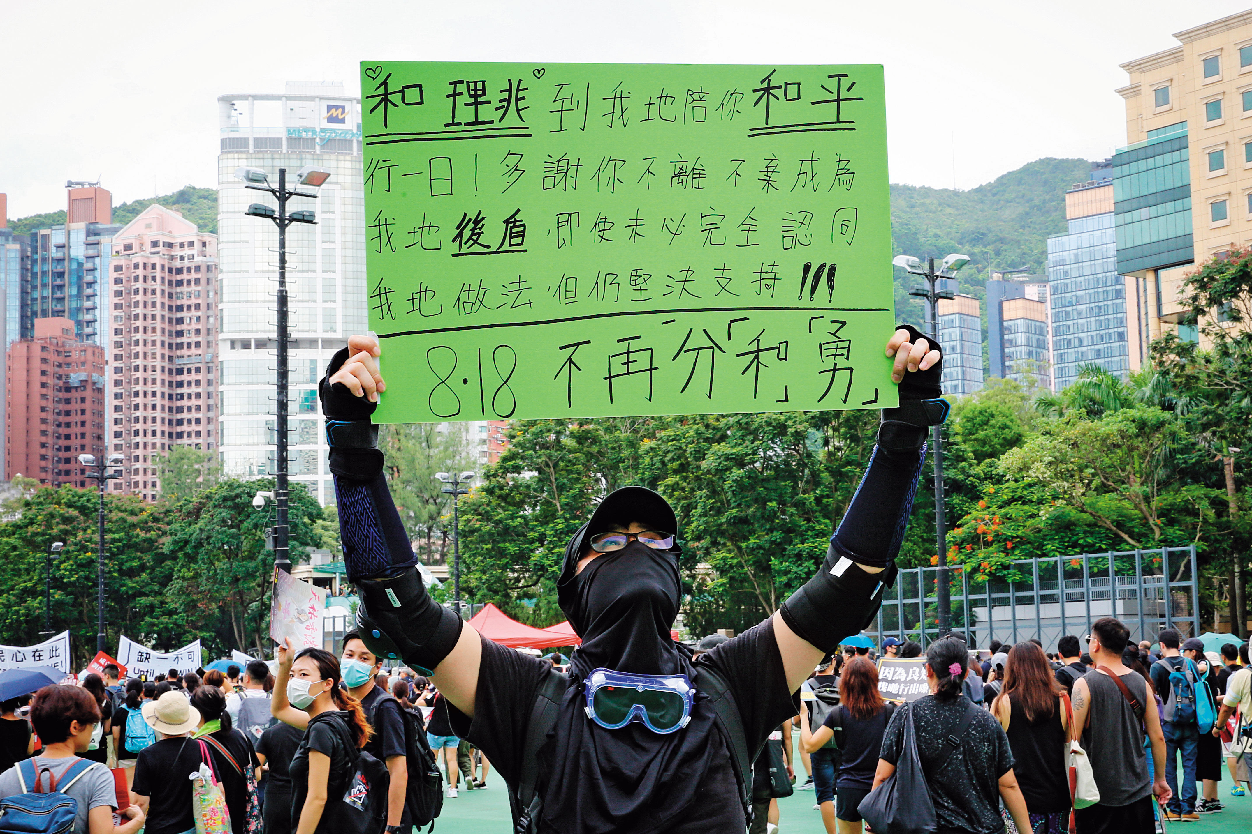 威爾（化名）高舉自備手寫看板，主張「818不再分『和』『勇』」，頂著高溫，在維園的一角苦站。