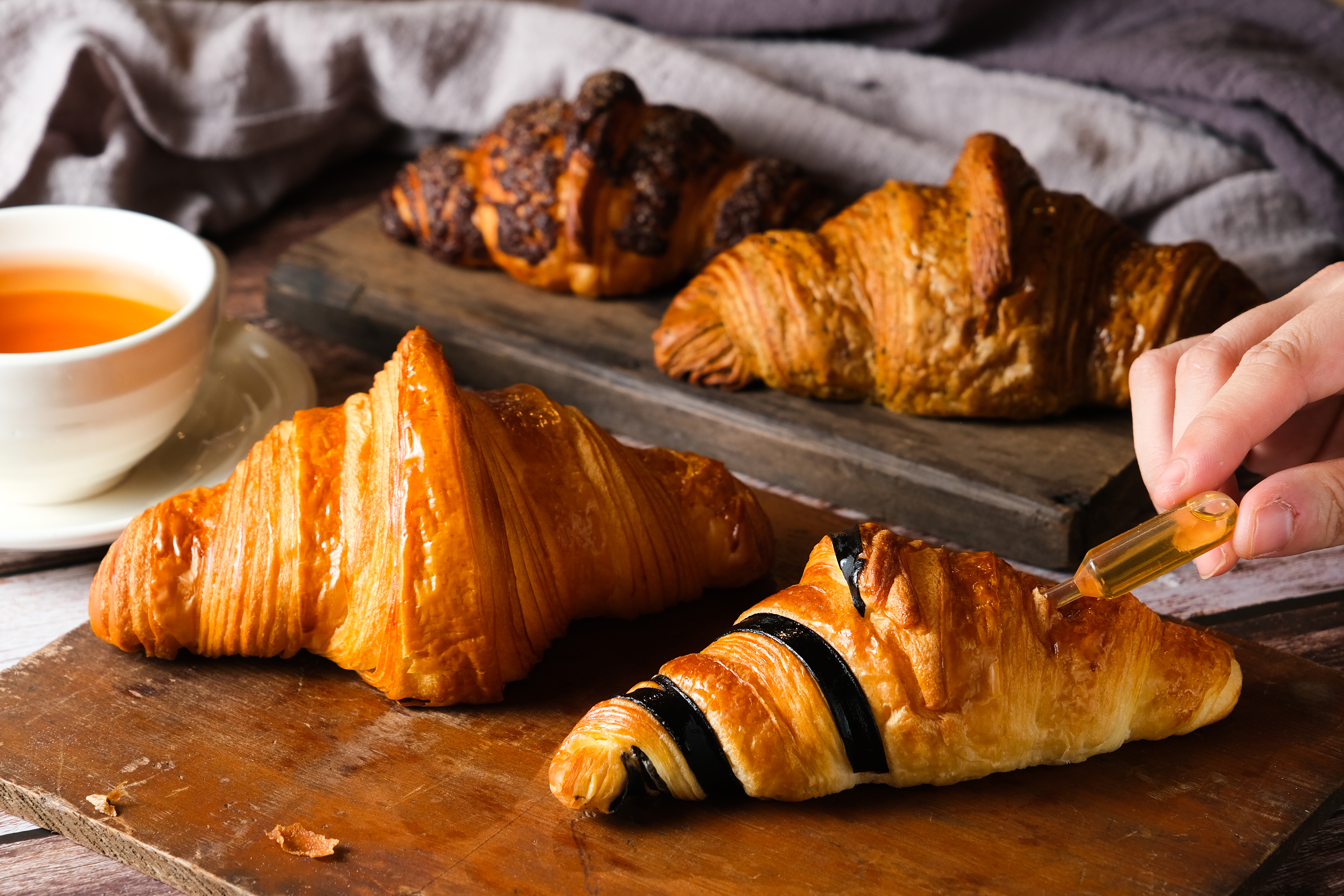 「蜜蜂可頌」（右前，65元／個，新光三越A11櫃位限定）可把蜂蜜擠在肚子裡，或淋在表面，蜜香濃稠。「原味可頌」（左前，50元／個）口感酥脆，奶香細緻。「台灣茶可頌」（右後，55元／個）茶香淡雅。「脆皮可可頌」（左後，60元／個）有濃郁巧克力醬。