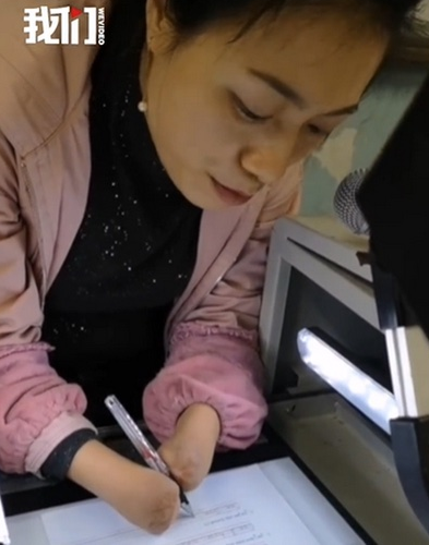 杜宣梅平時寫字需要用雙臂夾著筆,若要打開投影機則用雙臂夾著長棍開啟開關。(翻攝自新京報我們視頻)