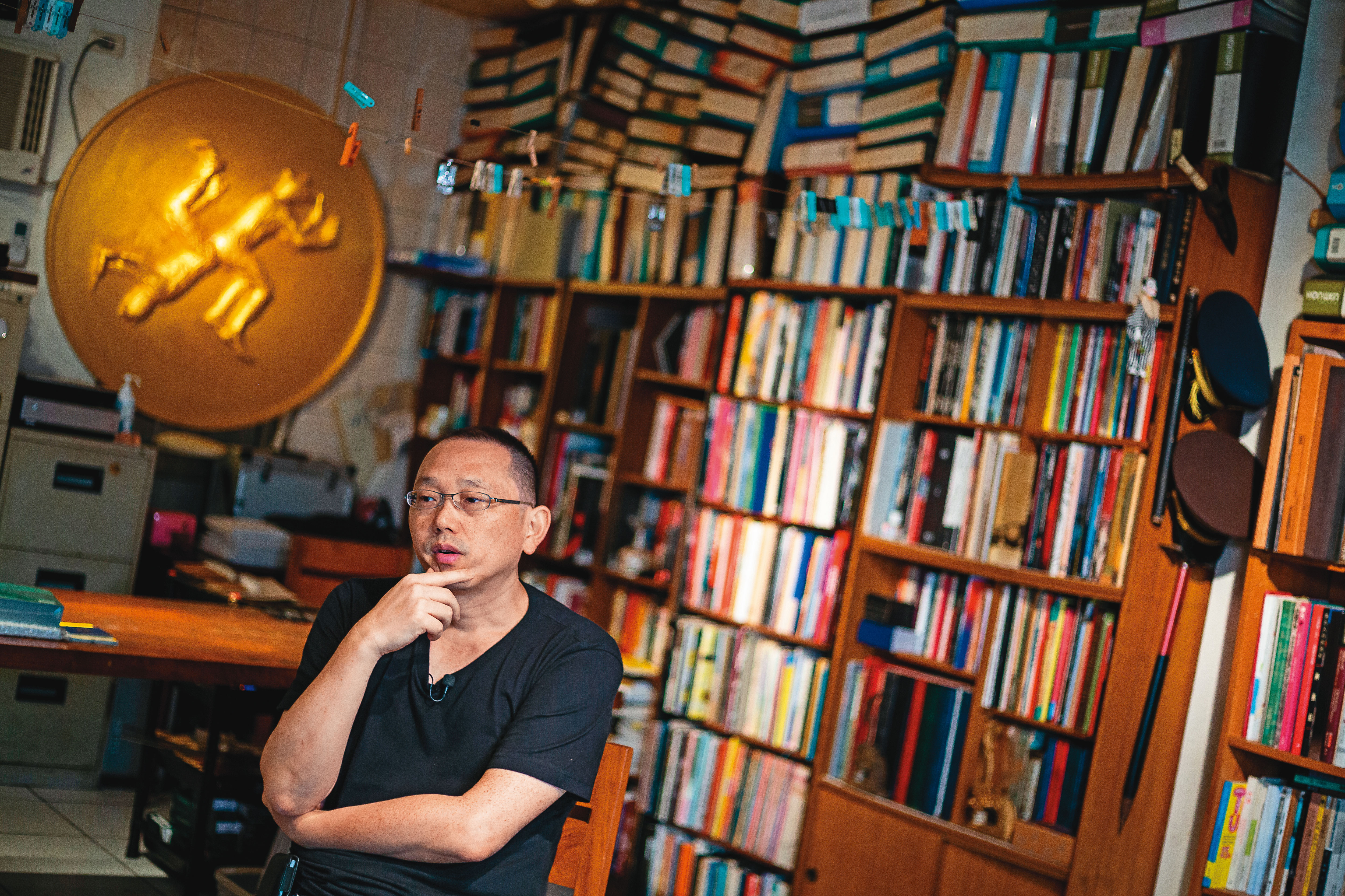 姚瑞中工作室（幻影堂）收藏了許多藝術書籍與展冊文獻，他身後的金色圓徽是他為今年大型個展《犬儒共和國》設計的國徽。