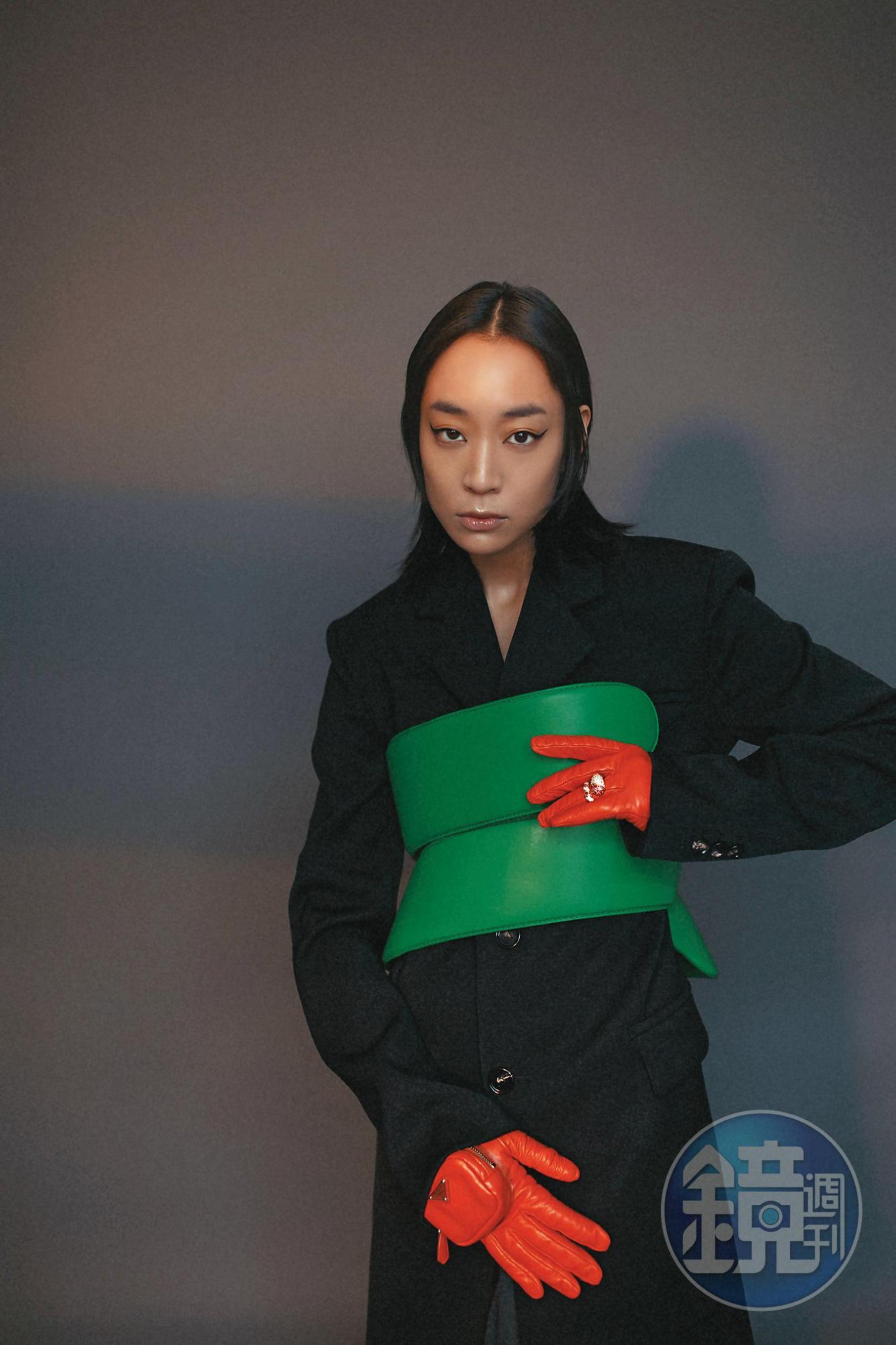 黑色單側雙口袋羊毛外套 NT$96,500 by BOTTEGA VENETA；鸚鵡綠螺旋繞身皮帶 NT$81,700 by BOTTEGA VENETA；紫紅白鋯石純銀鍍金戒指 NT$51,300 by BOTTEGA VENETA；橘色口袋皮革手套 NT$34,000 by PRADA。