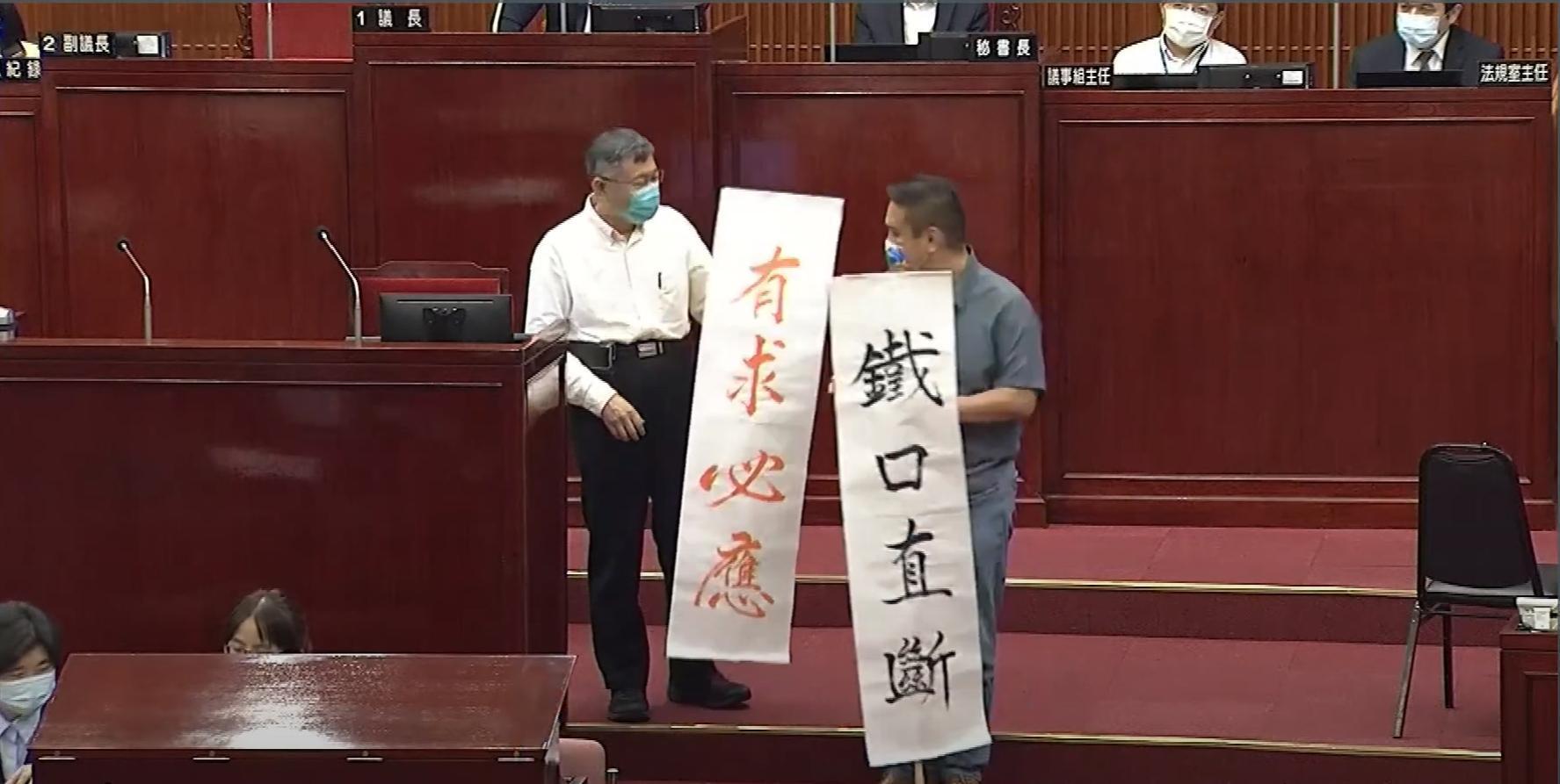 市議員陳重文（右）在質詢結束後，送給市長柯文哲（左）寫有「鐵口直斷」「有求必應」的2面旗幟。（翻攝自柯文哲YouTube頻道）