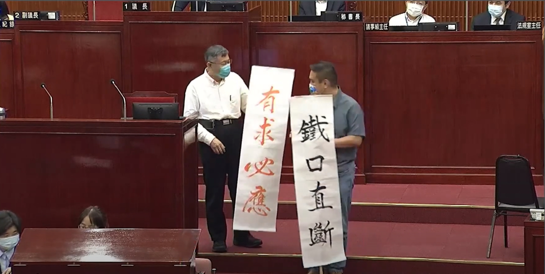 市議員陳重文（右）在質詢結束後，送給市長柯文哲（左）寫有「鐵口直斷」「有求必應」的2面旗幟。（翻攝自柯文哲YouTube頻道）