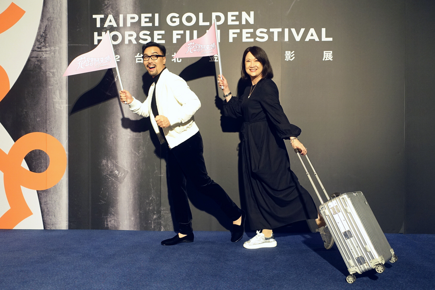 描述新加坡大媽獨自勇闖韓國旅行卻遭丟包，何書銘與洪慧芳呼應劇情，帶著導遊旗與旅行箱拍照。（采昌）