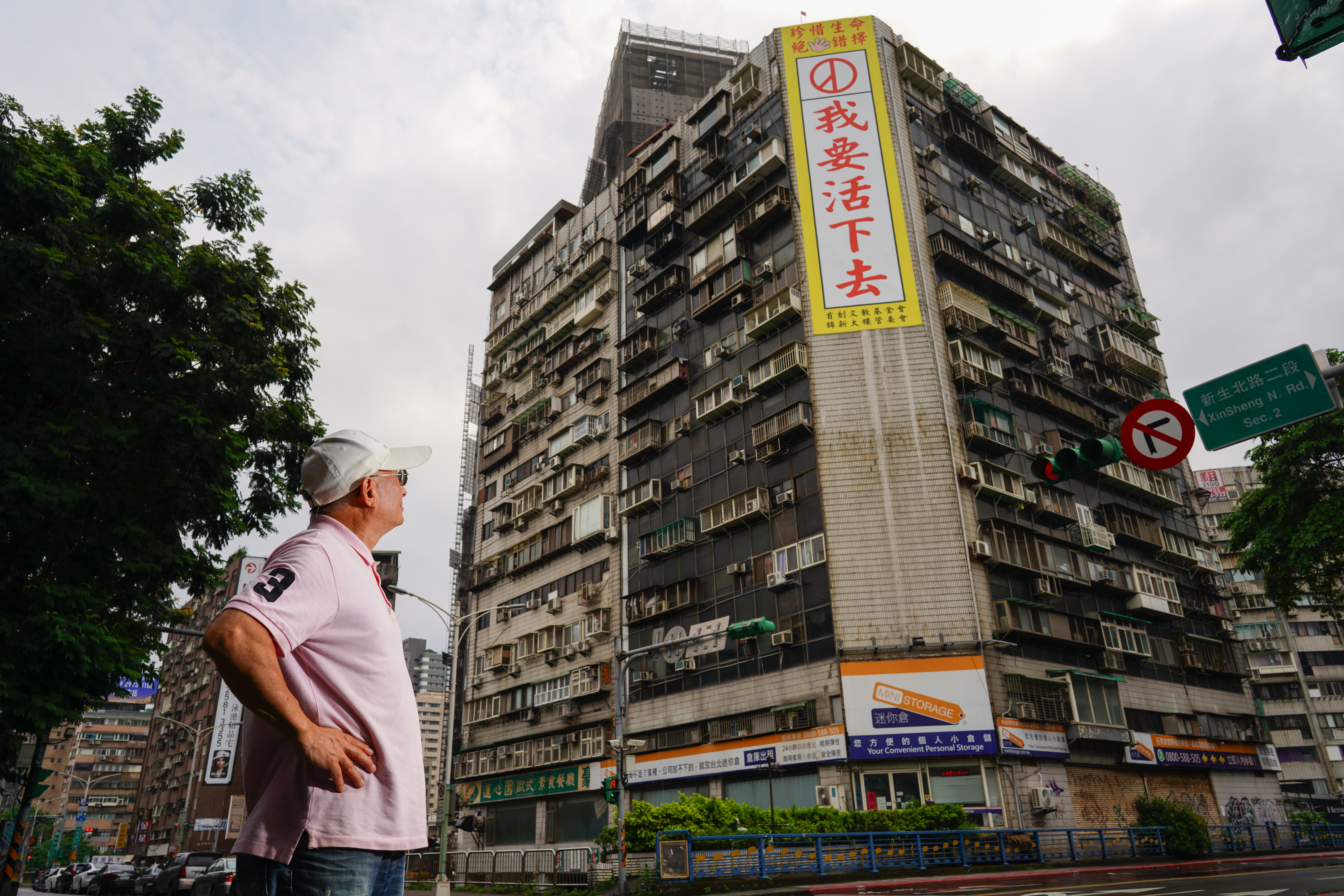 錦新大樓去年發生多起跳樓自殺事件。大樓管委會主委周朝陽於大樓外牆掛上廣告看板，期盼尋死者看到後能轉念。