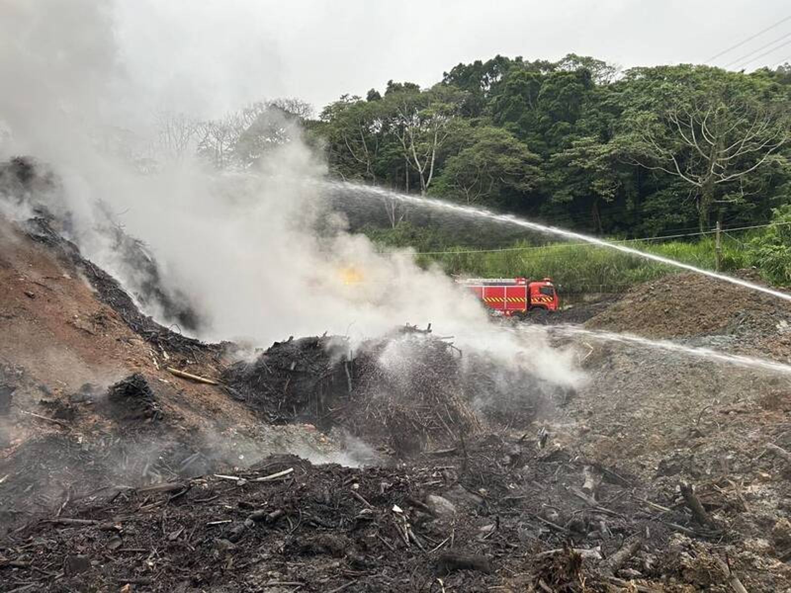 新竹縣府消防局邊開挖悶燒的廢木材、邊挖壕溝，注水後把起火的木材丟入浸泡以滅火。（消防局提供）