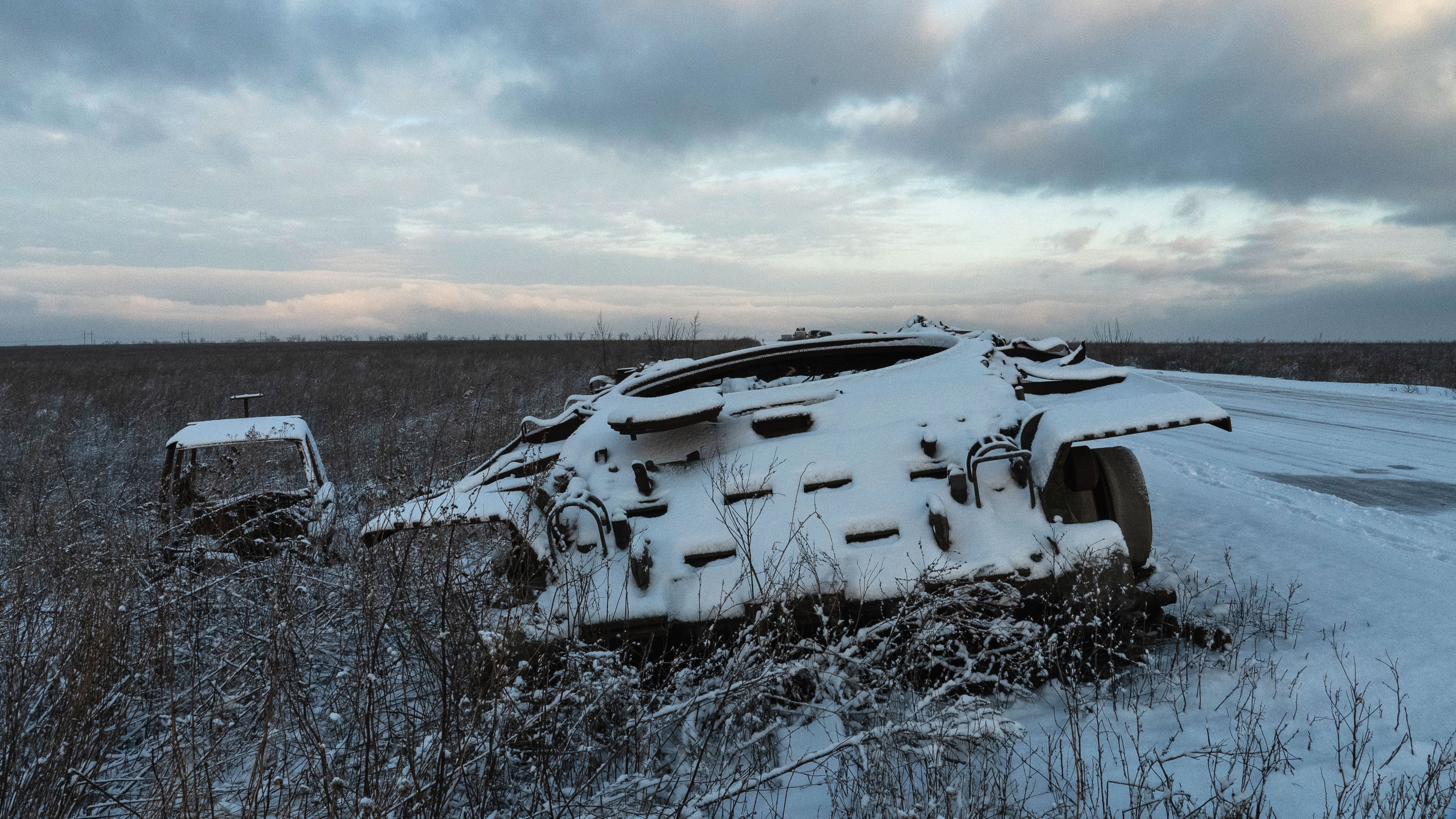 頓巴斯前線附近，白雪皚皚的路邊和鄰近的田野呈現出一片荒涼。一輛廢棄的坦克，履帶埋入雪中，像一位沉默的哨兵，見證了過去的衝突。在它旁邊，一輛遭燒毀的貨車使淒涼景象加倍，其受破壞的殘餘物銘蝕在冬景中。