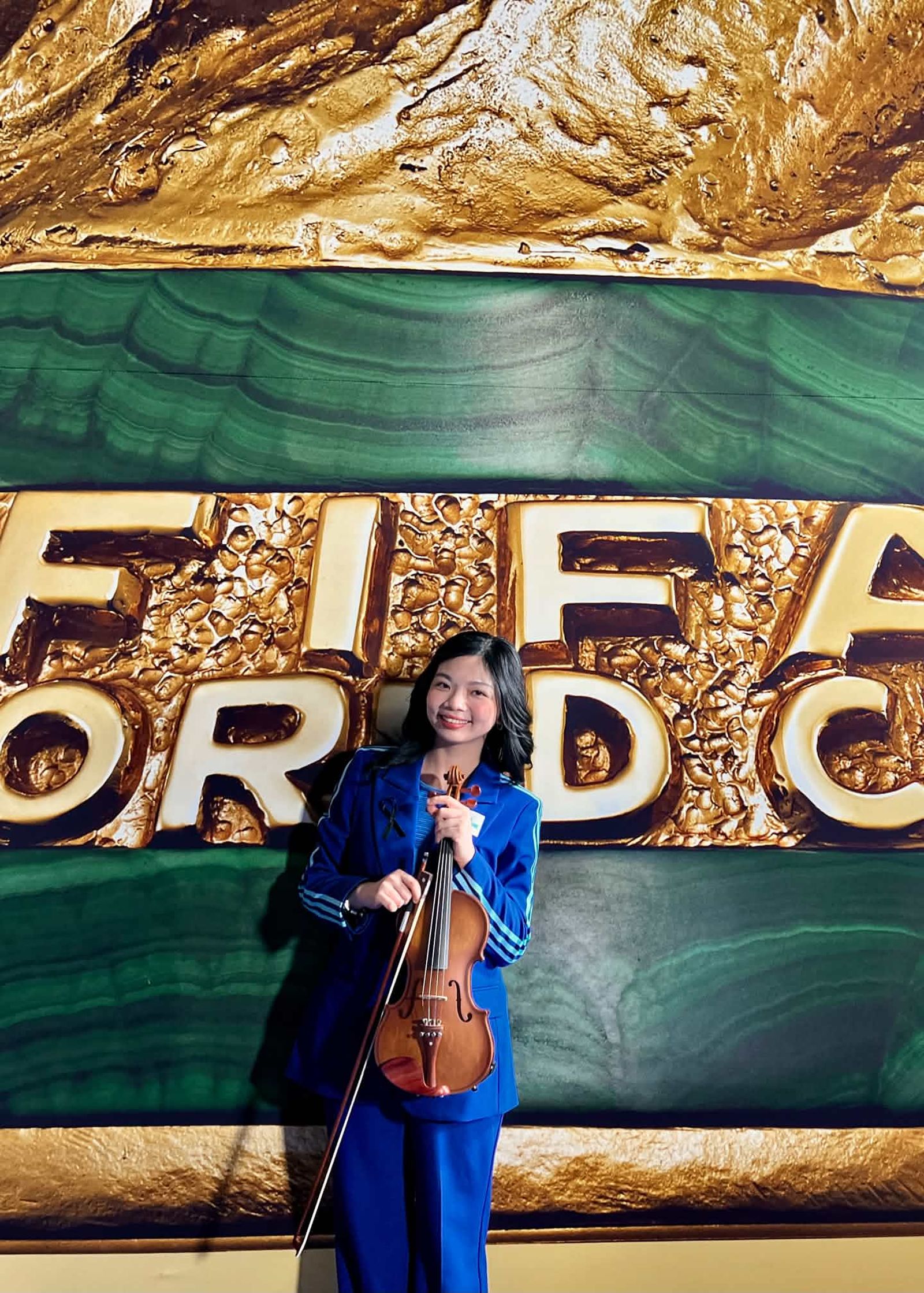 黃薏蓉拿著小提琴，在FIFA World Cup背板前留下紀念照片。（黃薏蓉提供）