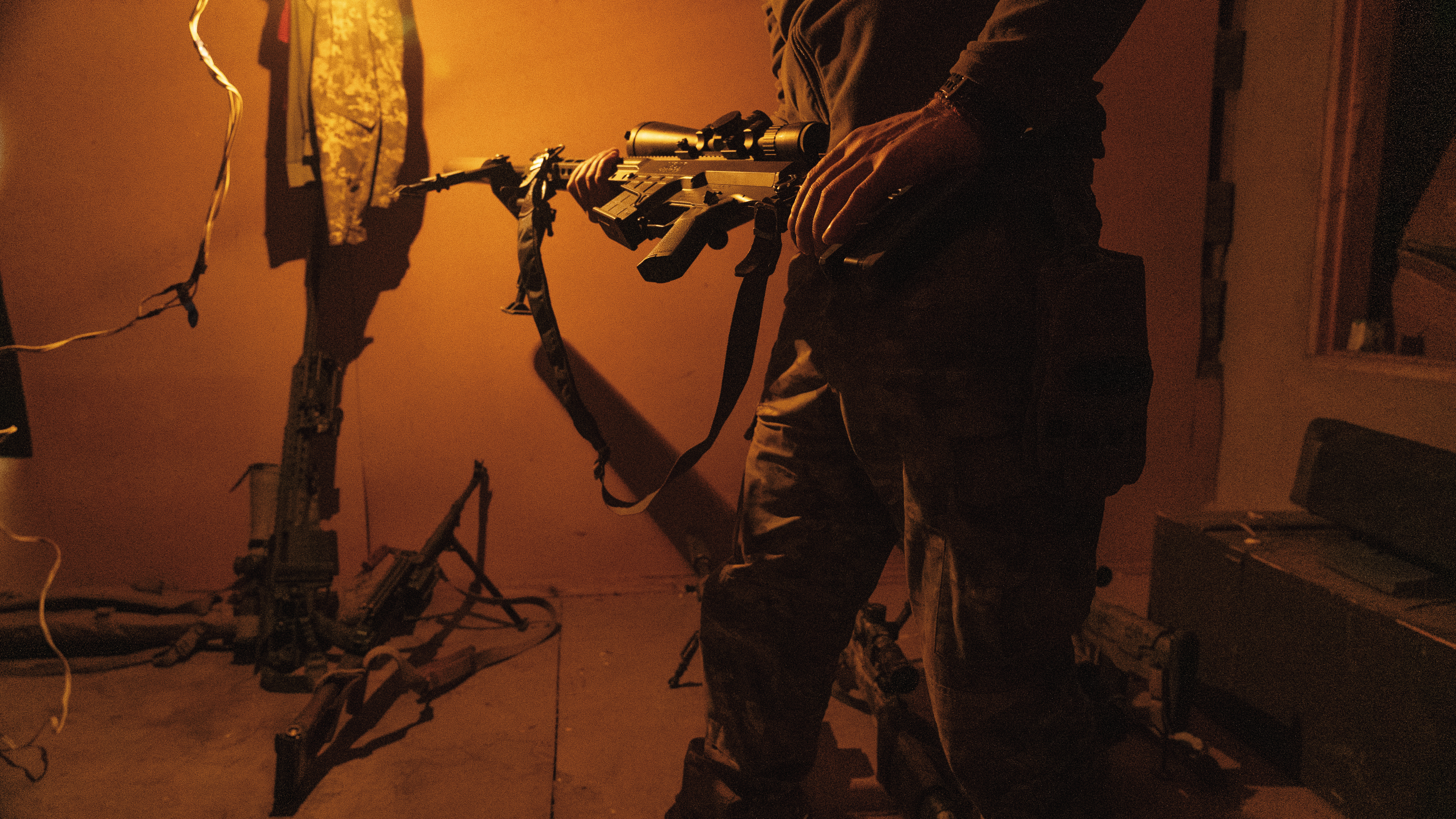 一名狙擊手沐浴在房間的橘色燈光裡，自信地從軍械庫中選出一把威力強大的步槍，他會在即將執行的任務中使用這把武器。